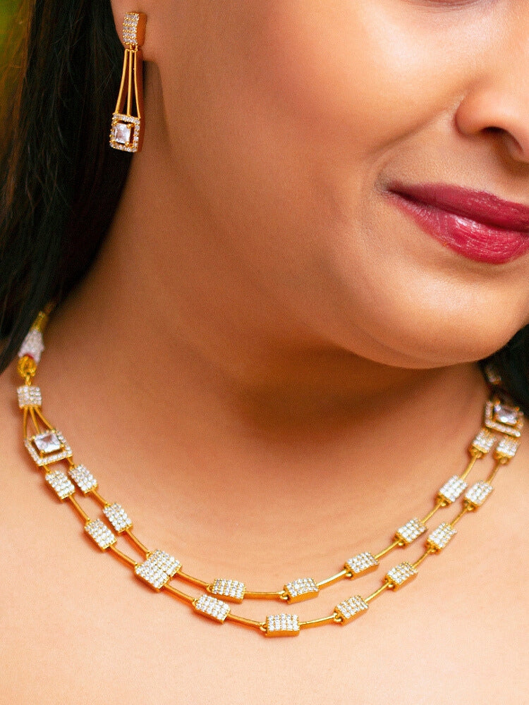 A woman wears the Diora White Necklace Set, a gold party jewelry set featuring a White 2 layer necklace set made with  American diamonds and White gemstones at both the corners, paired with matching earrings. Only her lower face and neck are visible.