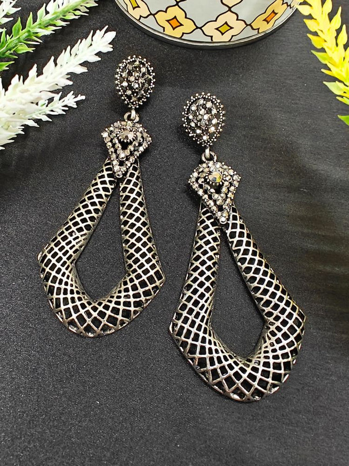 The Dipa Earrings are long oxidized earrings, gun metal grey pieces featuring intricate filigree and geometric patterns, showcased on a black surface with white and yellow foliage and a patterned container in the background.