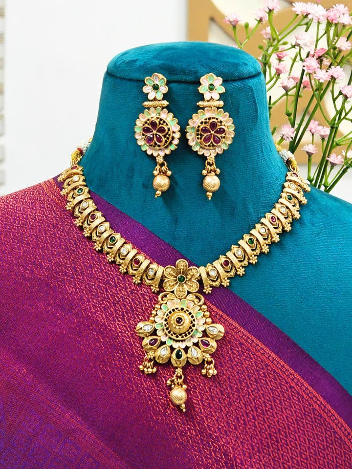A mannequin in a purple and teal sari showcases the Disha Necklace Set—an ornate gold necklace with matching floral earrings and colored stones, highlighting ethnic craftsmanship, set against a teal background with pink flowers.