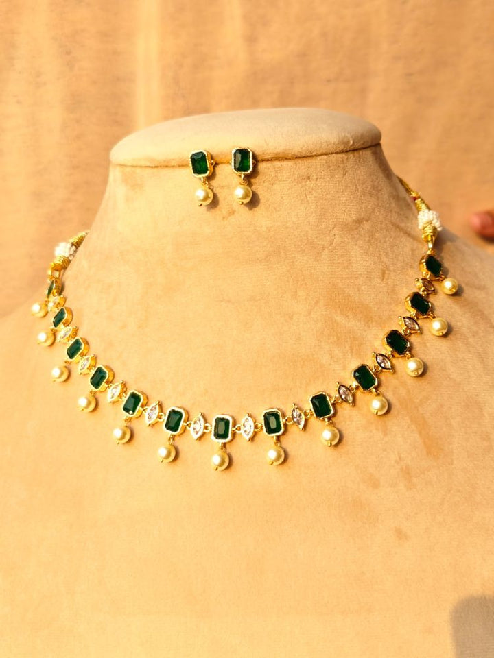 The Emerald Green Amelia Choker Necklace Set features colored stones and dangling pearl-like beads, displayed on a teal velvet stand, with matching stud earrings above and pink flowers beside the stand.