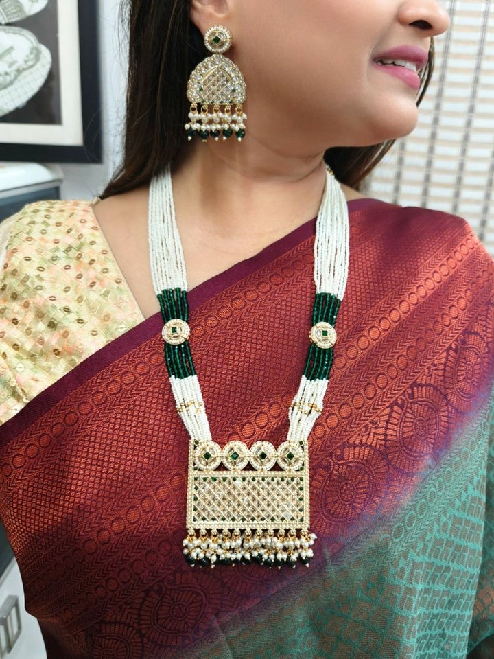A woman wearing the Falak Necklace Set—an ornate multiple layer pearl necklace, with a large rectangular pendant made with sparkling American Diamonds and hanging pearls at the bottom, and matching gold. American diamond and pearl earrings—paired with a maroon-green saree and gold blouse, smiles slightly.