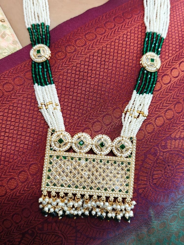 A woman wearing the Falak Necklace Set—an ornate multiple layer pearl necklace, with a large rectangular pendant made with sparkling American Diamonds and hanging pearls at the bottom, and matching gold. American diamond and pearl earrings—paired with a maroon-green saree and gold blouse, smiles slightly.