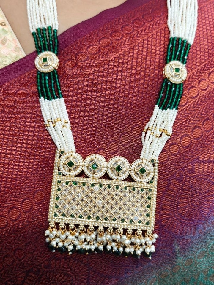 A woman wearing the Falak Necklace Set—an ornate multiple layer pearl necklace, with a large rectangular pendant made with sparkling American Diamonds and hanging pearls at the bottom, and matching gold. American diamond and pearl earrings—paired with a maroon-green saree and gold blouse, smiles slightly.