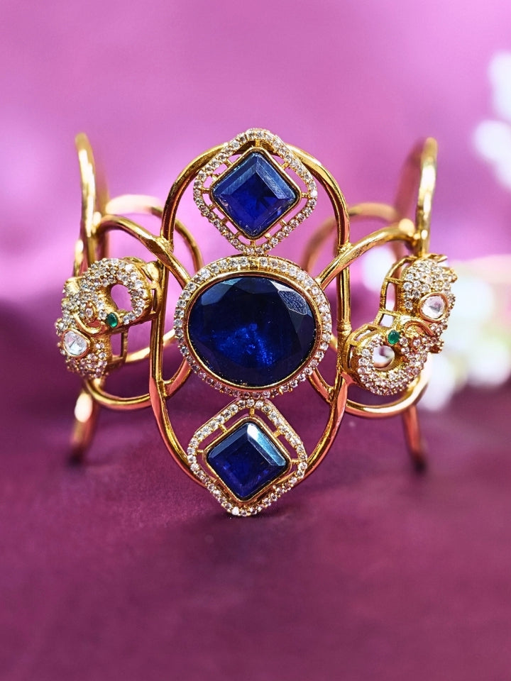 The Farah Navy Blue Bracelet (Adjustable) features intricate designs, three bold Navy Blue gemstones, and two elephant heads adorned with American diamonds —ideal for wedding ceremonies—shown on a purple fabric background.