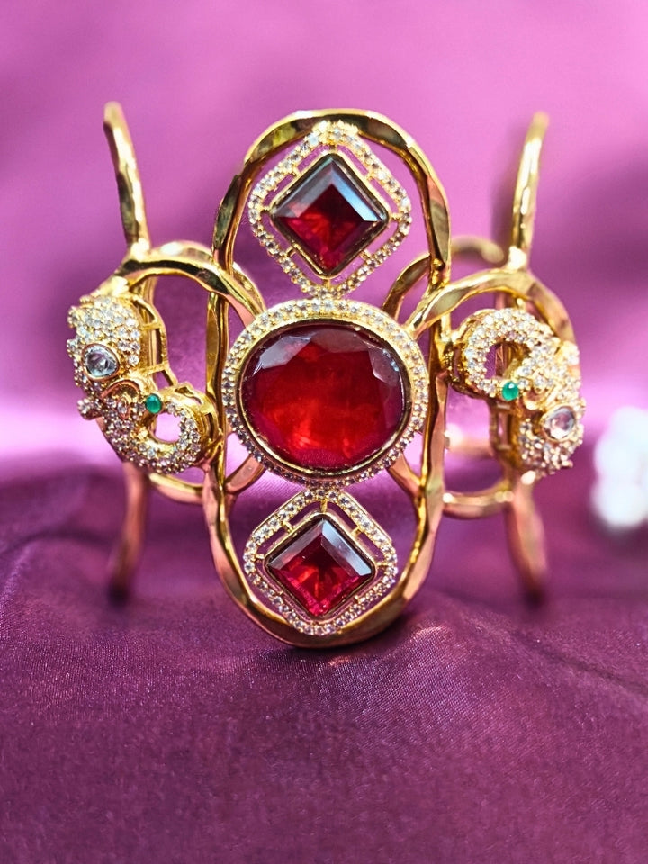 The Farah Red Bracelet (Adjustable) features intricate designs, three bold red gemstones, and two elephant heads adorned with American diamonds —ideal for wedding ceremonies—shown on a purple fabric background.