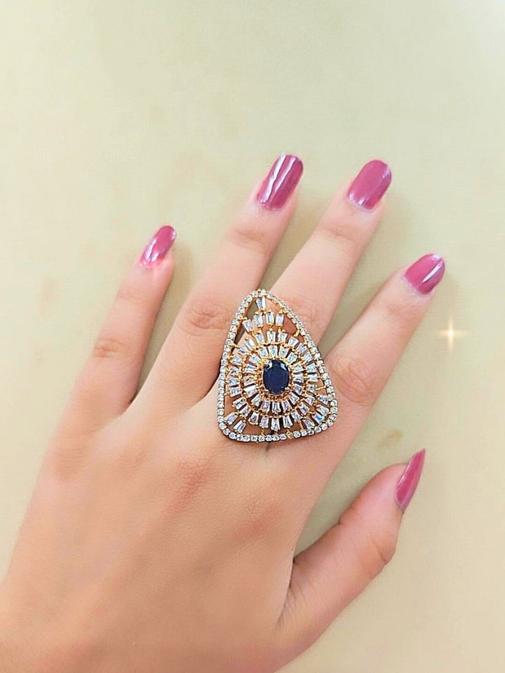 The Ferris Ring is a gold triangular adjustable ring featuring a central navy blue gemstone, surrounded by rows of sparkling American Diamonds, displayed on girls hand with pink nailpaint.  Perfect American Diamond cocktail ring. 