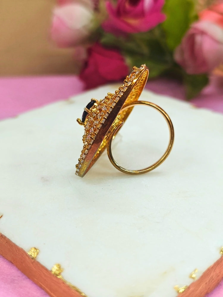 The Ferris Ring is a gold triangular adjustable ring featuring a central navy blue gemstone, surrounded by rows of sparkling American Diamonds, displayed on white marble with pink flowers in the background.  Perfect American Diamond cocktail ring. 