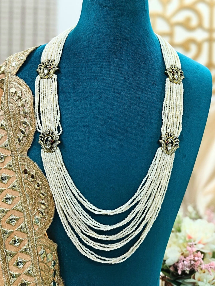 A mannequin showcases the Fiza Necklace, a multi-layered white pearl and beaded wedding piece with ornate gold and American Diamond clasps, styled over embroidered beige and gold fabric against a decorative backdrop.