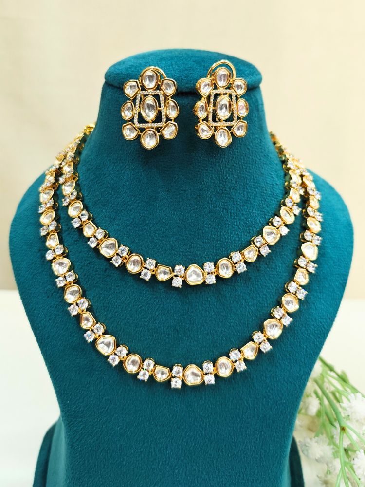 The Flora Necklace Set features a gold-tone, double-layered Tyani Kundan necklace and ornate stud earrings with round and teardrop Tyani Kundan stones, displayed on a teal jewelry stand—perfect for adding elegance to any outfit.