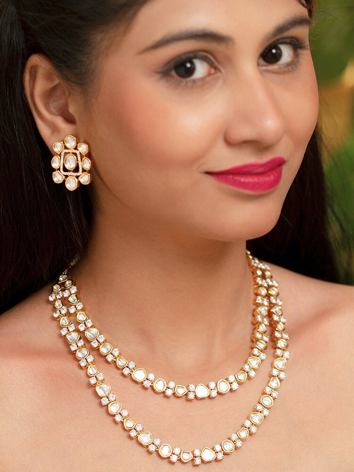 The Flora Necklace Set features a gold-tone, double-layered Tyani Kundan necklace and ornate stud earrings with round and teardrop Tyani Kundan stones, displayed on a teal jewelry stand—perfect for adding elegance to any outfit.