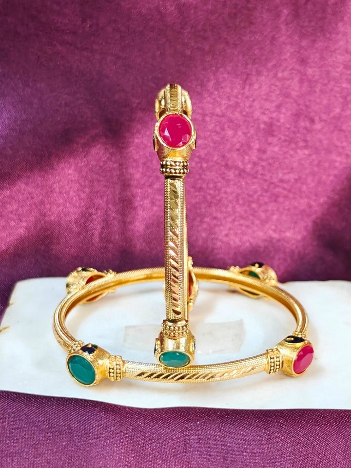 Close-up of an ornate traditional (Gold Polish) Bangles set with Red and Green Colored Stone. Festive celebration look enhanced with artificial Colored Stone bangles set. Festive Navratri outfit paired with stacked imitation Colored Stone bangles.