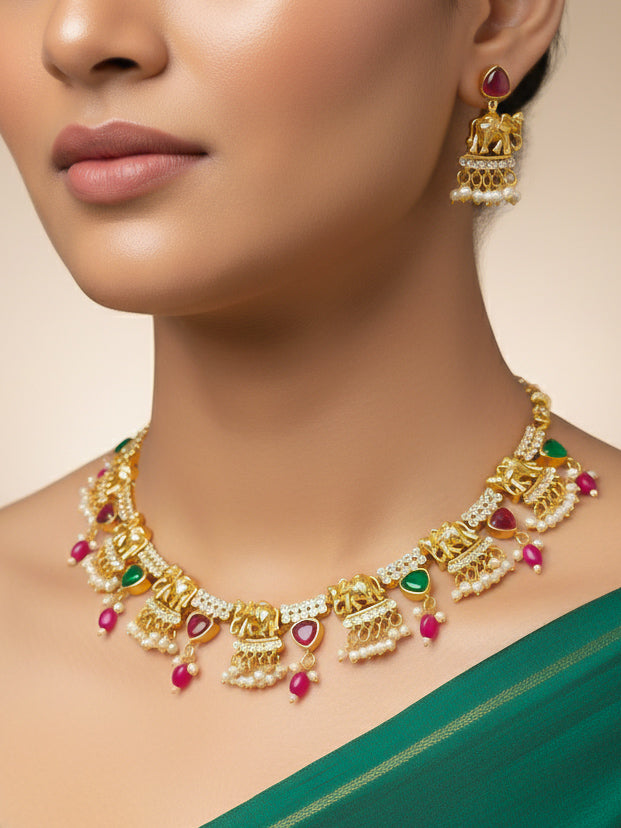 A woman wearing the Gajkesari Necklace Set featuring a traditional gold Collar Pink & Green necklace and matching earrings with pink and green colored stones, golden elephants and American Diamond. Close-up of a handcrafted temple imitation necklace styled with a silk saree. Woman wearing a temple-inspired artificial jewelry set for festive celebrations.