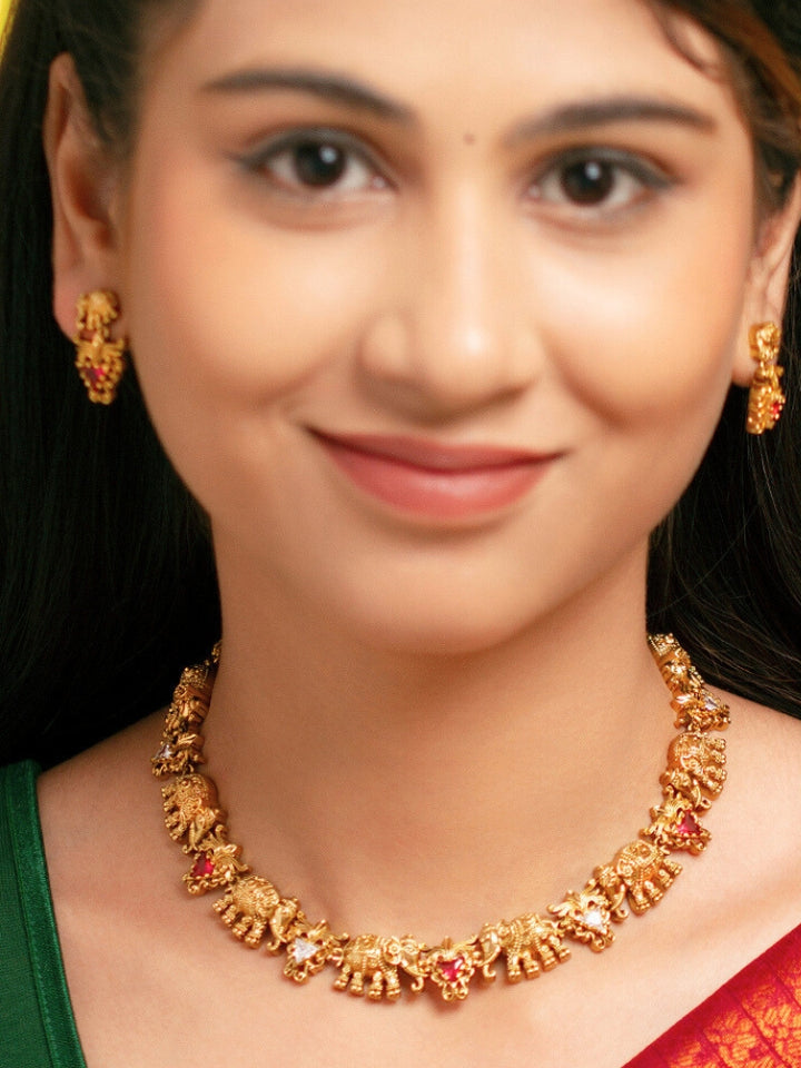A woman smiles in a green and red saree, adorned with the Gajvani Red & White Necklace Set—gold necklace and earrings featuring intricate elephant designs with red & White colored stones. Great simple elegant temple jewelry necklace set for festive occasions 