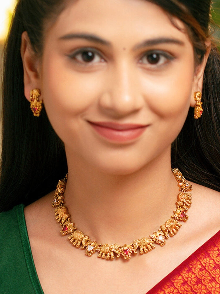 A woman smiles in a green and red saree, adorned with the Gajvani Red & White Necklace Set—gold necklace and earrings featuring intricate elephant designs with red & White colored stones. Great simple elegant temple jewelry necklace set for festive occasions 