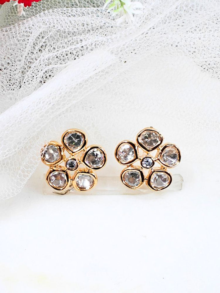 A pair of Gardenia tyaani kundan Earrings, featuring gold floral designs with five clear american diamond petals and a central gemstone, displayed on white textured fabric.