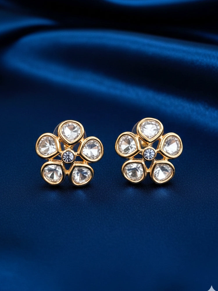 A pair of Gardenia tyaani kundan Earrings, featuring gold floral designs with five clear american diamond petals and a central gemstone, displayed on white textured fabric.