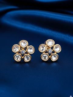 A pair of Gardenia tyaani kundan Earrings, featuring gold floral designs with five clear american diamond petals and a central gemstone, displayed on white textured fabric.