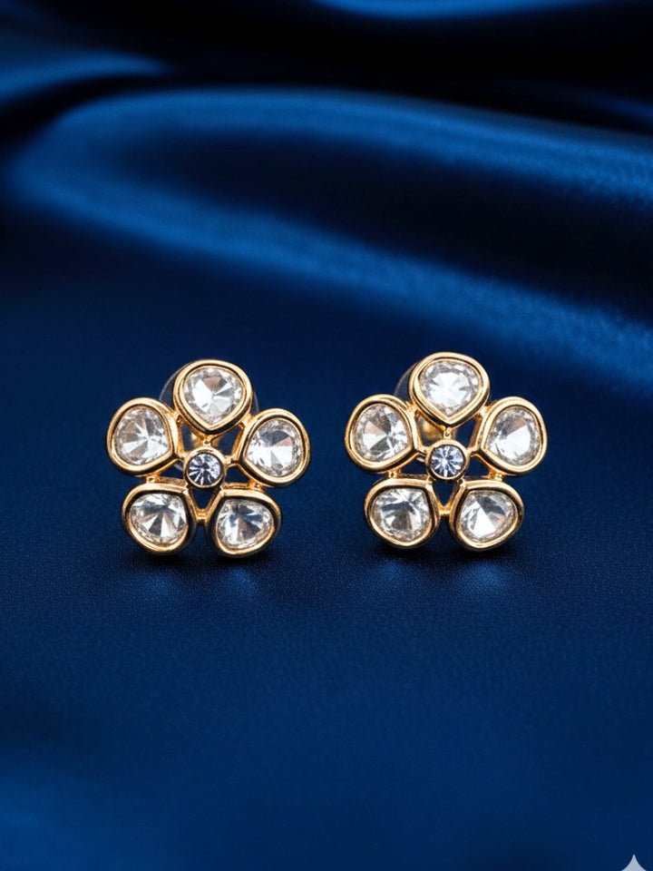 A pair of Gardenia tyaani kundan Earrings, featuring gold floral designs with five clear american diamond petals and a central gemstone, displayed on white textured fabric.
