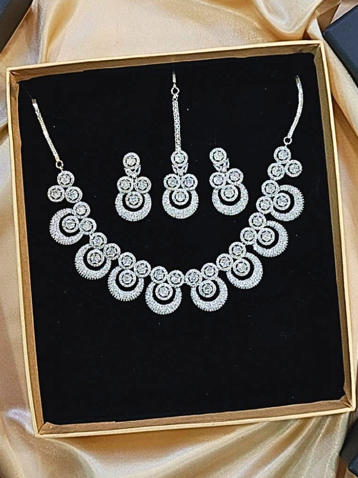 The Genevieve Necklace Set includes a sparkling silver necklace with matching earrings and a pendant, all adorned with circular American Diamonds and elegantly presented on black velvet against a gold satin fabric background.