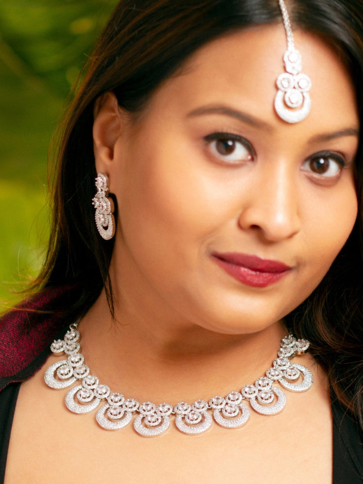 The Genevieve Necklace Set includes a sparkling silver necklace with matching earrings and a pendant, all adorned with circular American Diamonds and elegantly presented on black velvet against a gold satin fabric background.