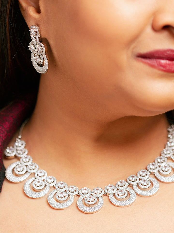 The Genevieve Necklace Set includes a sparkling silver necklace with matching earrings and a pendant, all adorned with circular American Diamonds and elegantly presented on black velvet against a gold satin fabric background.
