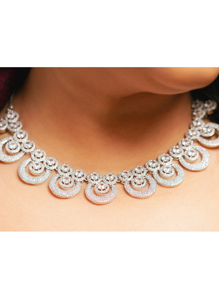 The Genevieve Necklace Set includes a sparkling silver necklace with matching earrings and a pendant, all adorned with circular American Diamonds and elegantly presented on black velvet against a gold satin fabric background.