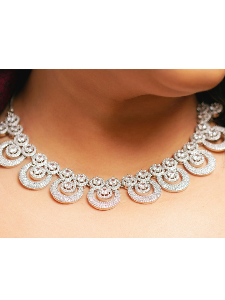 The Genevieve Necklace Set includes a sparkling silver necklace with matching earrings and a pendant, all adorned with circular American Diamonds and elegantly presented on black velvet against a gold satin fabric background.