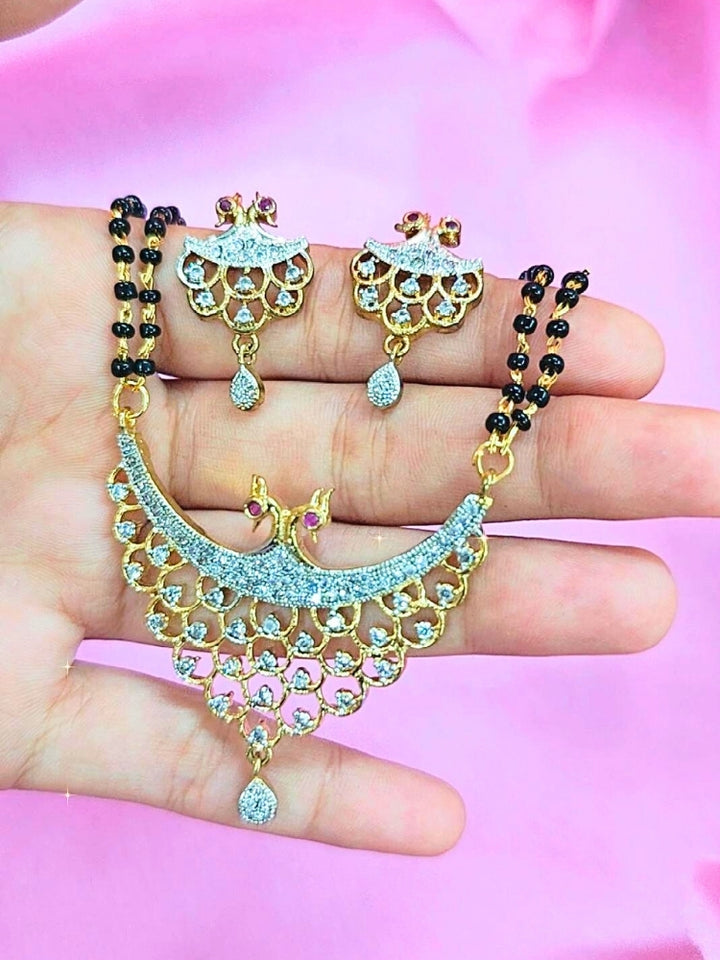 A hand holds the Gita Mangalsutra—a gold necklace with black mangalsutra beads and matching earrings, pendant featuring sparkling American Diamonds—set against a soft pink background, perfect for your wedding ceremony dress. Diamond mangalsutra. Modern mangalsutra. 