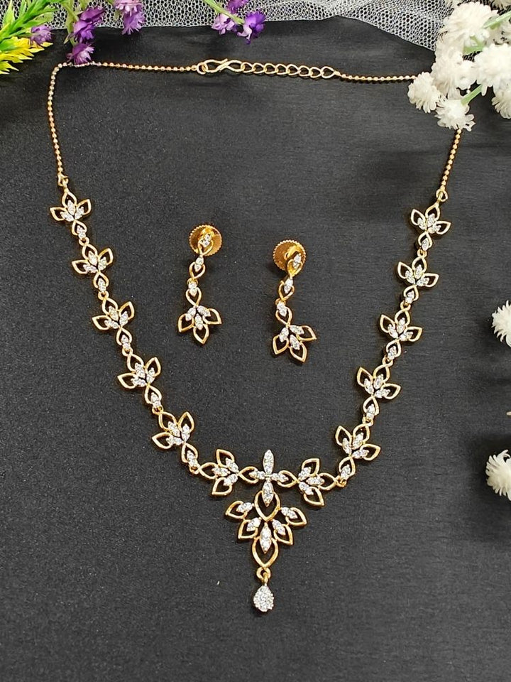 The Gold Madison Necklace Set features sparkling American diamonds along with intricate gold floral designs with matching earrings, beautifully displayed on a black surface accented by flowers—perfect as elegant wedding jewelry.