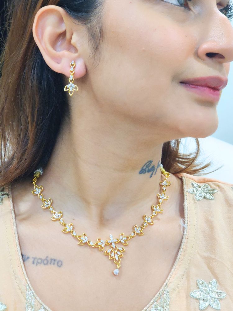 The Gold Madison Necklace Set features sparkling American diamonds along with intricate gold floral designs with matching earrings, beautifully displayed on a girl looking on the side—perfect as elegant wedding jewelry.