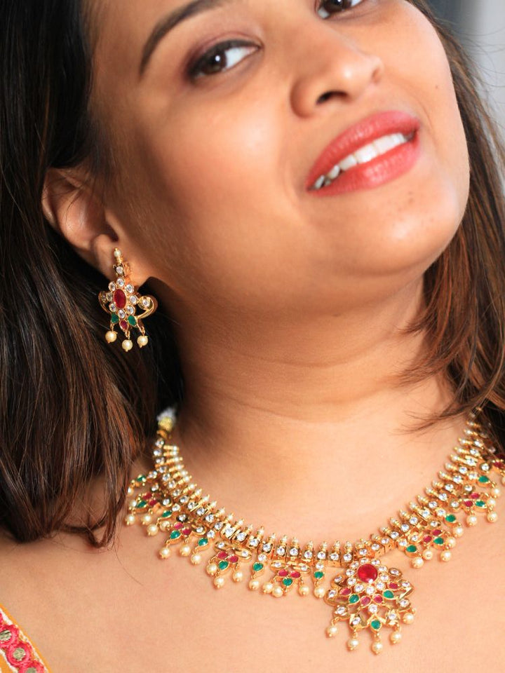 A woman smiles, wearing the Gold Priyamvada Necklace Set featuring matching earrings adorned with red and green gemstones and pearls. This intricate set of traditional wedding jewelry exudes timeless elegance.