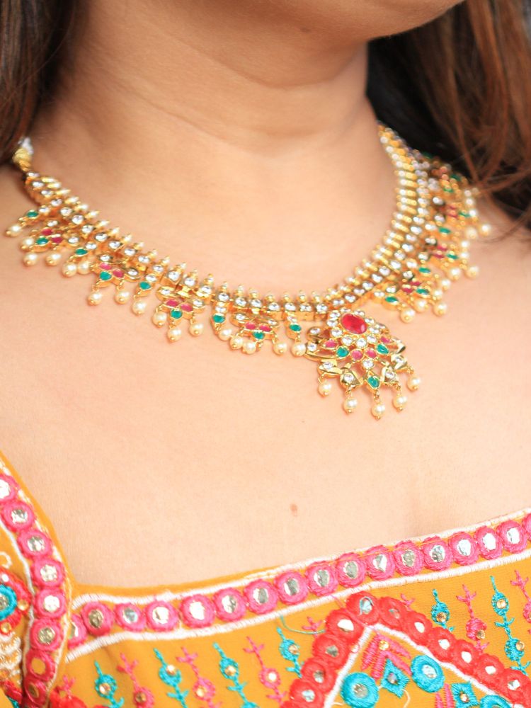A woman smiles, wearing the Gold Priyamvada Necklace Set featuring matching earrings adorned with red and green gemstones and pearls. This intricate set of traditional wedding jewelry exudes timeless elegance.