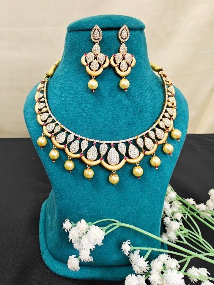 The Gold Sakshi Necklace Set, featuring American diamonds designed in a teardrop shape in the middle of the length of the necklace with intricate detailing, and dangling gold colored pearls with matching earrings, is displayed on a teal stand accented with white artificial flowers at the base.