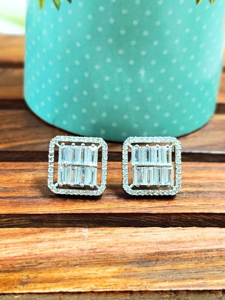 The Goldie Silver Studs are square-shaped earrings with American Diamonds in a baguette pattern, displayed on wood before a turquoise polka dot container for a chic look.