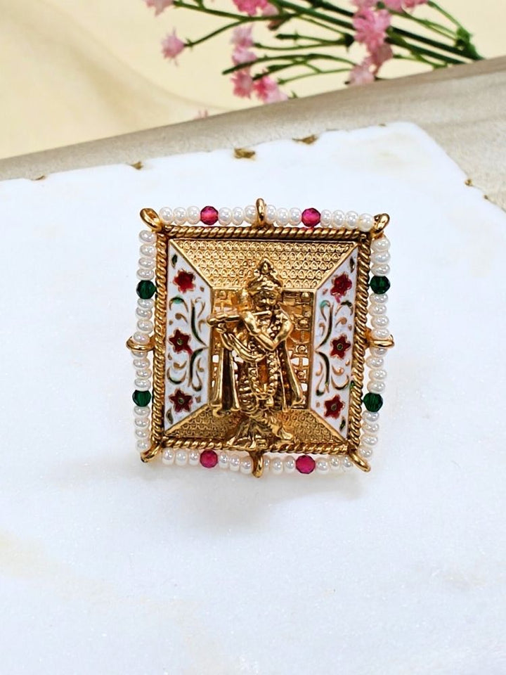 The Gopal Ring is a square, gold-toned adjustable ring featuring Krishna at the center, colorful enamel floral designs, pearl accents, and red-green bead details—an exquisite ethnic jewelry piece shown with pink flowers.