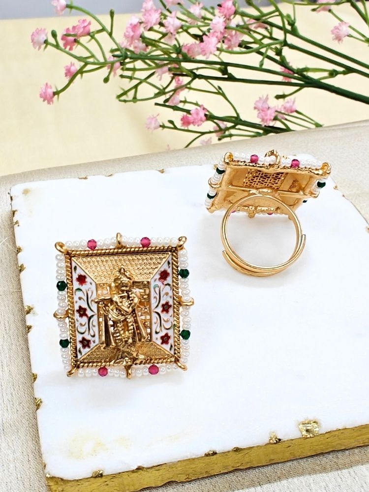 The Gopal Ring is a square, gold-toned adjustable ring featuring Krishna at the center, colorful enamel floral designs, pearl accents, and red-green bead details—an exquisite ethnic jewelry piece shown with pink flowers.