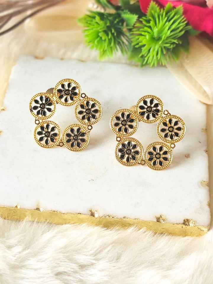 The Gracie Earrings are gold-toned alloy circular stud earrings adorned with black floral Meenakari work, shown on a white marble slab with gold edges, and surrounded by green and red artificial foliage.