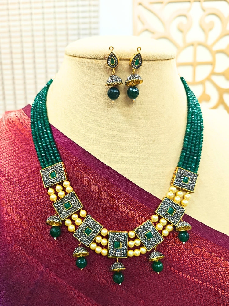 The Green Anahita Necklace Set features a necklace with multi-strand green crystals, with squares filled with black crystals and green colored stone at the center, connecting multiple squared with ivory colored pearls, and small jhumki style hangings at the bottom. It comes along with matching earirngs. Shown on a mannequin in a red and purple silk saree.