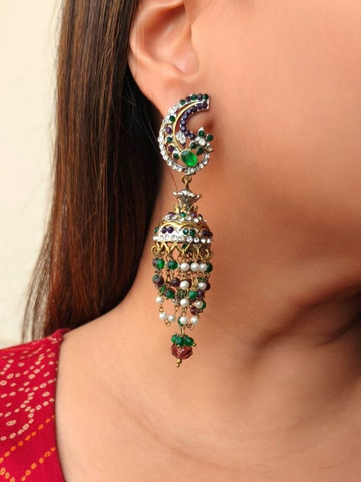 A close-up of a woman with straight brown hair wearing the Green Charu Earrings—large, ornate Victorian-style earrings adorned with intricate designs, colorful beads, dangling elements, and striking red, green, and gold accents.