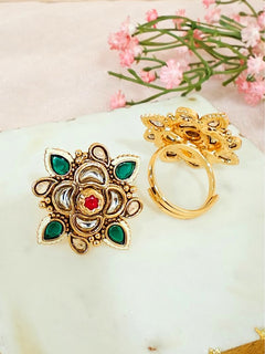 The Green Chhavi Ring features ornate gold brass ring with floral designs and green, white, and red colored stones. Adjustable and elegant, it’s shown on marble with pink flowers in the background—perfect for elevating any look.