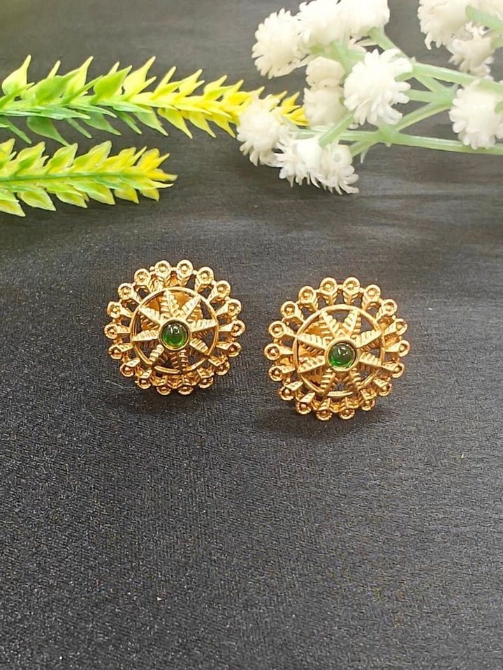 The Green Gehna Earrings are traditional round gold stud earrings with intricate designs and a central Emerald-colored stone, displayed on a dark surface with white and yellow artificial flowers in the background.