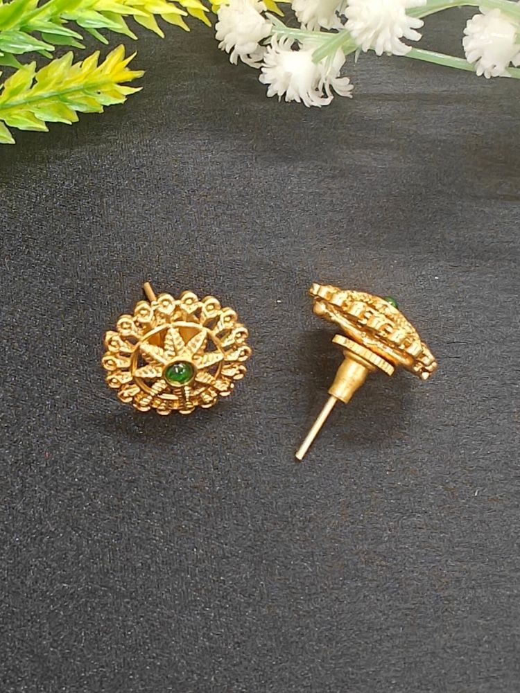 The Green Gehna Earrings are traditional round gold stud earrings with intricate designs and a central Emerald-colored stone, displayed on a dark surface with white and yellow artificial flowers in the background.