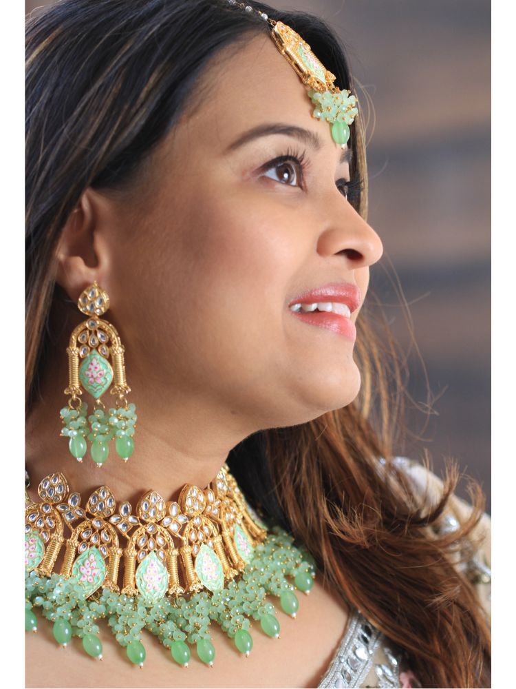 A woman with long dark hair wears the Green Jodha Necklace Set, featuring a heavy green Kundan Meena necklace with kundan, matching ornate earrings, and maangtikka. She smiles softly at the camera. Perfect meenakari kundan necklace for weddings. 