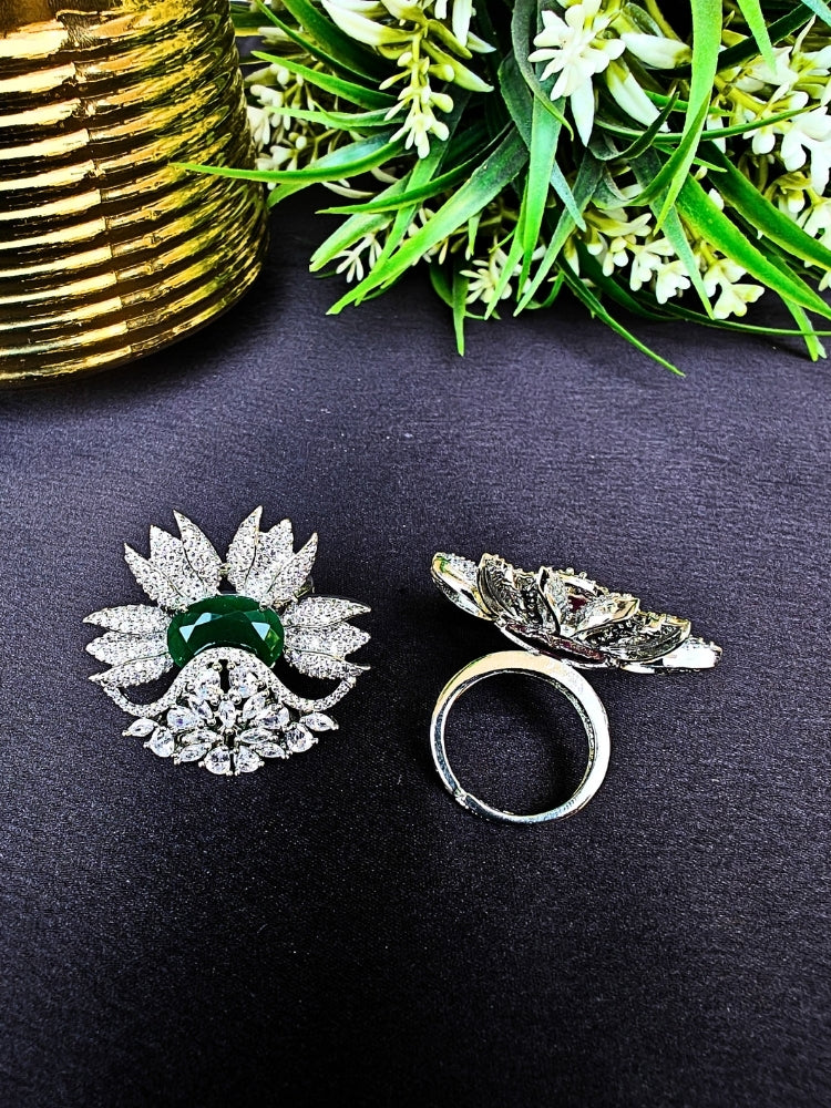 Two ornate, flower-shaped cocktail rings rest on a dark surface; one is the Green Megan Ring, featuring Emerald Green stones and sparkling American Diamond accents. The other ring is shown from behind, with green foliage and a gold object in the background.