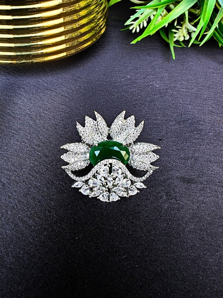 flower-shaped cocktail rings rest on a dark surface; this is the Green Megan Ring, featuring Emerald Green stones and sparkling American Diamond accents. The ring is shown with green foliage and a gold object in the background.