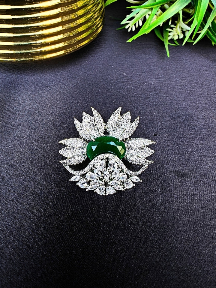 flower-shaped cocktail rings rest on a dark surface; this is the Green Megan Ring, featuring Emerald Green stones and sparkling American Diamond accents. The ring is shown with green foliage and a gold object in the background.