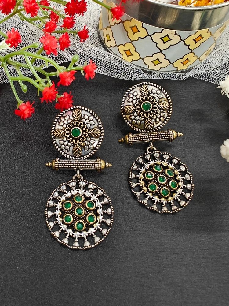 The Green Sheetal Earrings feature intricate circular brass designs with embedded American diamonds, offering an antique oxidized look. Displayed with red flowers and a patterned container, they’re perfect for a wedding ceremony. Perfect to wear on navratri. 