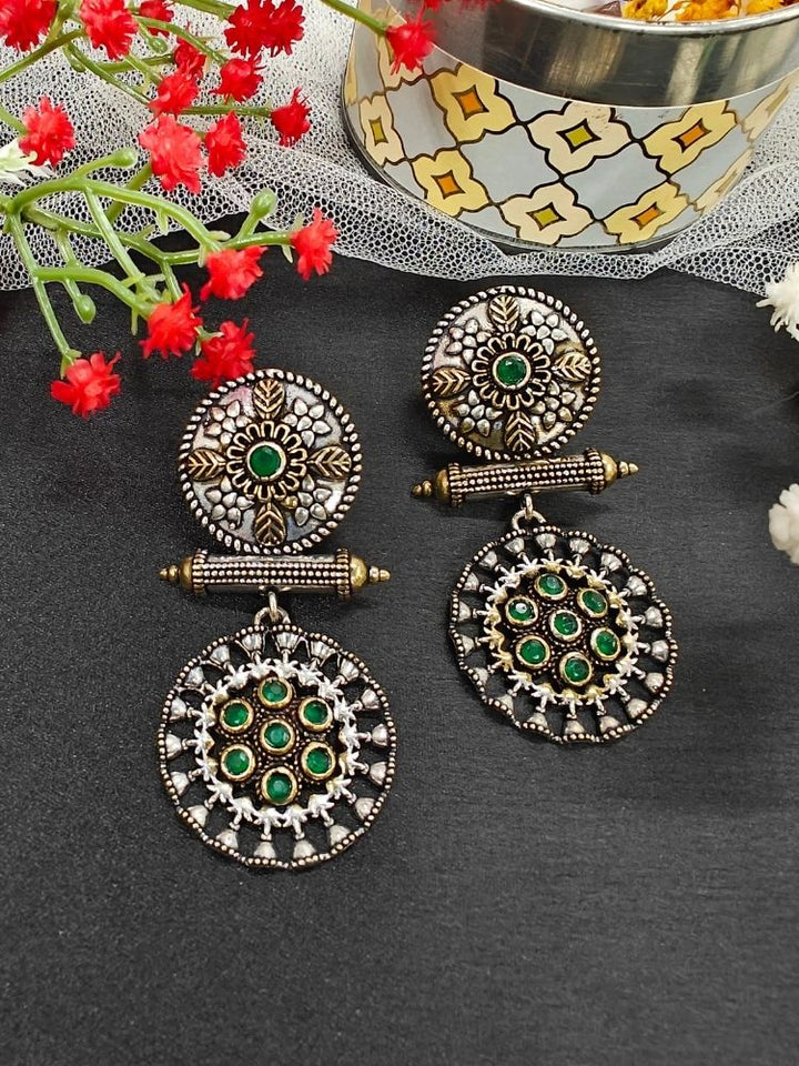 The Green Sheetal Earrings feature intricate circular brass designs with embedded American diamonds, offering an antique oxidized look. Displayed with red flowers and a patterned container, they’re perfect for a wedding ceremony. Perfect to wear on navratri. 
