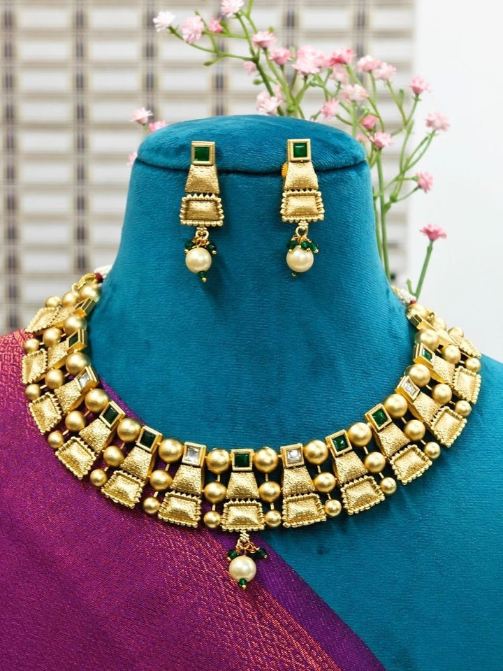 A mannequin displays the Green Sheetla Necklace Set, featuring a brass gold necklace and matching earrings with geometric patterns and pearl details, against a backdrop of pink flowers and a window.