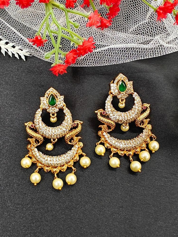 The Green Zarina Earrings are ornate gold and American Diamond earrings with pearl dangles and intricate detailing, displayed on a black surface with white netting and red flowers. Perfect to complement your wedding ceremony dress.